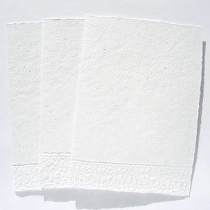 3 weiße Klappkarten aus handgeschöpftem Papier mit geprägtem Spitzenrand an der unteren Seite von paperuli liegen im Hochformat fächerförmig auf dem Tisch.