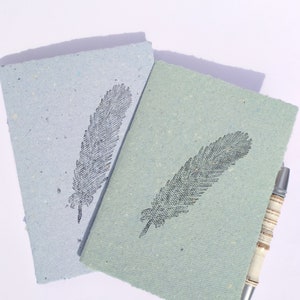ein blaues und ein grünes kleines Notizheft im Postkarten format aus handgeschöpftem Papier von paperuli mit schwarzem Motivstempel Feder, daneben ein Kugelschreiber zur Veranschaulichung der Größe