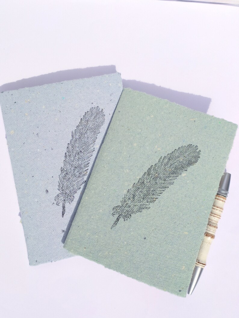 ein blaues und ein grünes kleines Notizheft im Postkarten format aus handgeschöpftem Papier von paperuli mit schwarzem Motivstempel Feder, daneben ein Kugelschreiber zur Veranschaulichung der Größe