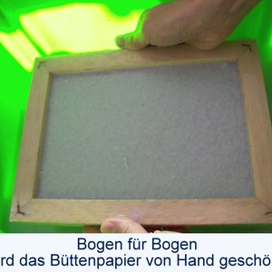 Zwei Hände heben einen nassen hölzernen Schöpfrahmen aus einer hellgrünen Plastikwanne. Auf dem Rahmen  liegt ein weißer nasser Bogen Papier. Unter dem Foto steht der Text: "Bogen für Bogen wird das Büttenpapier von Hand geschöpft".