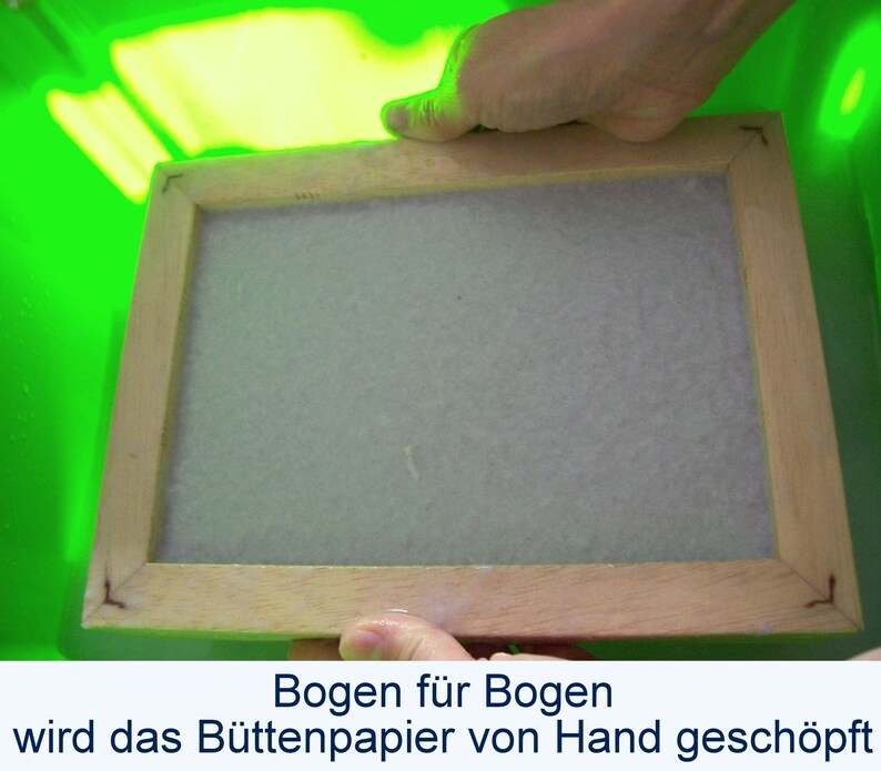 Zwei Hände heben einen nassen hölzernen Schöpfrahmen aus einer hellgrünen Plastikwanne. Auf dem Rahmen  liegt ein weißer nasser Bogen Papier. Unter dem Foto steht der Text: "Bogen für Bogen wird das Büttenpapier von Hand geschöpft".