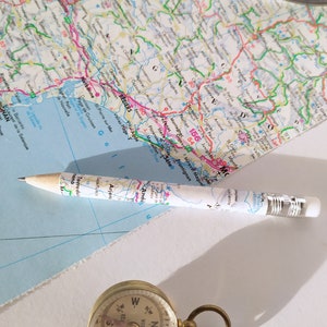 Può includere: Un primo piano di una bussola con una custodia dorata e un quadrante bianco. La bussola è appoggiata su una superficie bianca. Una matita con una mappa avvolta intorno è appoggiata su una mappa della Francia. La mappa è blu, verde e rosa.