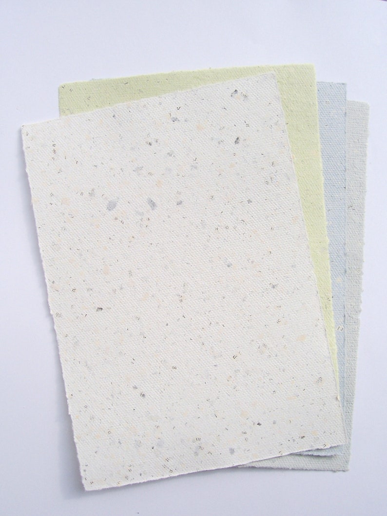 4 Bogen Recycling-Büttenpapier von paperuli mit deutlich sichtbaren Buchseiten-Fragmenten im Papier, in 4 Farben: creme, hellgrün, hellblau, hellgrau, liegen fächerförmig auf dem Tisch.