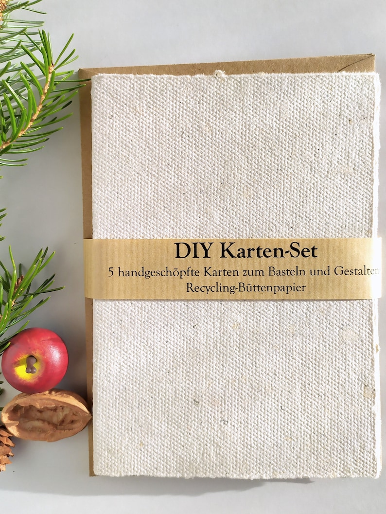 naturfarbene Büttenkarten mit Kraftpapier-Umschlägen, mit einer Info-Banderole aus Kraftpapier umwickelt, von paperuli. Am linken Bildrand weihnachtliche Deko aus Tanne, Zier-Apfel und Nussschalen.