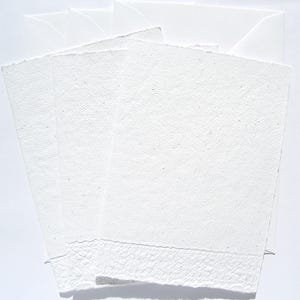 3 weiße Klappkarten aus handgeschöpftem Papier mit geprägtem Spitzenrand an der unteren Seite von paperuli liegen fächerförmig im Hochformat auf 3 weißen Briefumschlägen auf dem Tisch.