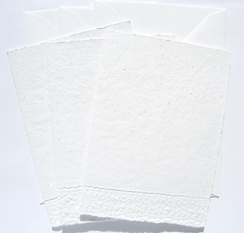 3 weiße Klappkarten aus handgeschöpftem Papier mit geprägtem Spitzenrand an der unteren Seite von paperuli liegen fächerförmig im Hochformat auf 3 weißen Briefumschlägen auf dem Tisch.