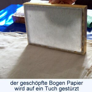 Eine Hand stellt einen Schöpfrahmen senkrecht auf ein Tuch, damit der darauf befindliche frisch geschöpfte Bogen Papier abgegautscht werden kann. Unter dem Foto steht der Text: "der geschöpfte Bogen Papier wird auf ein Tuch gestürzt".