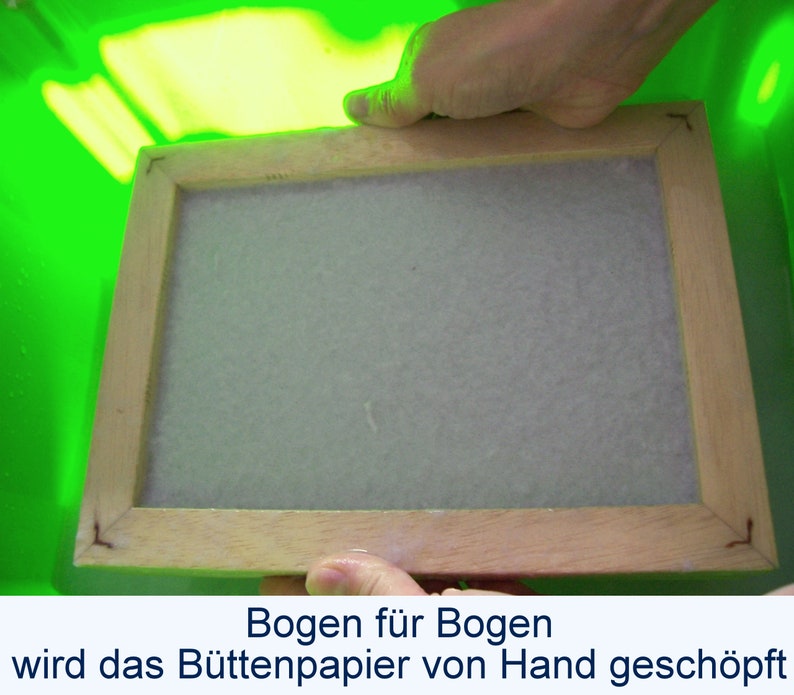 Zwei Hände heben einen nassen hölzernen Schöpfrahmen aus einer hellgrünen Plastikwanne. Auf dem Rahmen liegt ein nasser weißer Bogen Papier. Unter dem Foto steht der Text: „Bogen für Bogen wird das Büttenpapier von Hand geschöpft“.