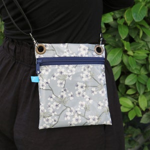 Puede incluir: Un pequeño bolso bandolera con estampado floral con flores blancas sobre un fondo azul claro. El bolso tiene cierre de cremallera y dos anillos metálicos en la parte superior.