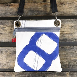 Puede incluir: Bolso bandolera de lona blanca con un diseño de ocho azul y blanco. El bolso tiene una cremallera y una correa gris.