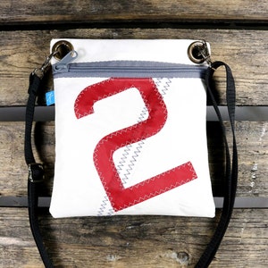 Puede incluir: Un bolso bandolera de lona blanca con un número "2" rojo en la parte delantera. El bolso tiene una cremallera y una correa negra ajustable.