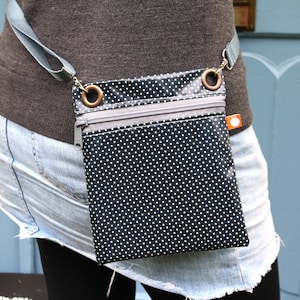 Puede incluir: Un pequeño bolso bandolera de lunares negros y blancos con una correa gris y herrajes plateados. El bolso tiene dos compartimentos con cremallera.