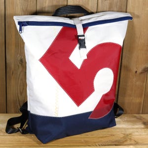Op de afbeelding: Witte en marineblauwe rugzak met een groot rood ontwerp. De rugzak heeft een zwart handvat en bandjes, een marineblauwe rits en een witte gesp. De tas is gemaakt van duurzaam materiaal.