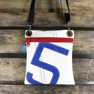 Puede incluir: Un bolso bandolera de lona blanca con un número "5" azul y una cremallera roja. El bolso tiene una correa ajustable negra.