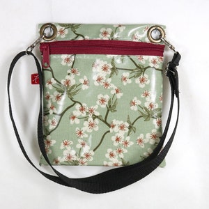 Puede incluir: Un pequeño bolso bandolera de estampado floral verde y blanco con una correa negra ajustable y una cremallera roja.