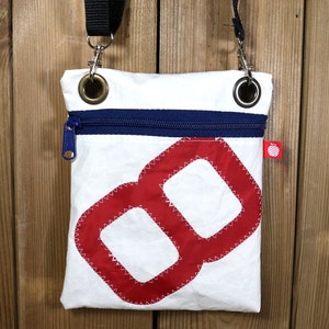 Puede incluir: Bolso bandolera de lona blanca con un diseño náutico rojo y blanco. El bolso tiene una cremallera y una correa negra ajustable.