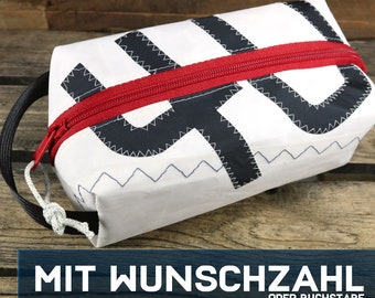 May include: White zippered toiletry bag with a red zipper and black and white geometric design. The bag has a handle and is made from recycled sailcloth.  MIT WUNSCHZAHL ODER BUCHSTABE