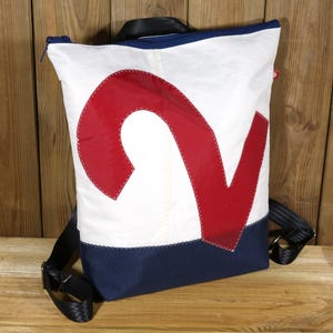 Puede incluir: Mochila blanca con un gran número rojo "2" aplicado. La mochila tiene una base azul marino y correas negras ajustables. La cremallera es negra y el fondo es una superficie de madera.