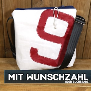 Könnte beinhalten: Weiße Umhängetasche mit einer großen roten Neun. Die Tasche hat einen schwarzen, verstellbaren Riemen und einen blauen Reißverschluss. Der Text "MIT WUNSCHZAHL ODER BUCHSTABE" befindet sich am unteren Bildrand.