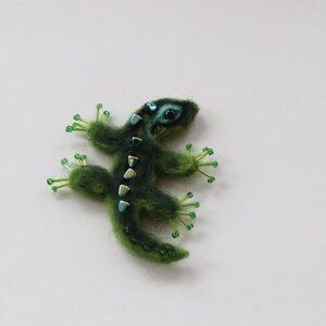 May include: A handmade, green lizard brooch with a textured, fuzzy appearance. The lizard has small, light green beaded feet and a row of iridescent, rectangular accents down its back. The eyes are a bright turquoise color.