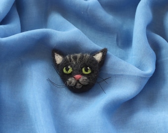 Può includere: Spilla a forma di gatto fatta a mano in feltro. La testa del gatto è nera con occhi verdi, naso rosa e punte delle orecchie bianche. La spilla è posta su uno sfondo di tessuto blu.