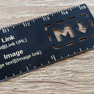 Peut inclure: Une règle noire avec des marques dorées et une découpe en forme de lettre "M" avec une flèche vers le bas. La règle est graduée de 10 à 15. Le texte "Link [Link text](Link URL)" et "Image [Image text](Image link)" est imprimé sur la règle.