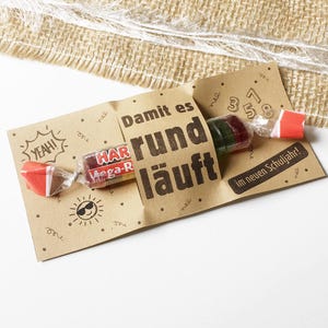 May include: A brown paper gift tag with the text "Damit es rund läuft" and a wrapped candy. The tag has a sun graphic and the word "YEAH!". A burlap ribbon is in the background.