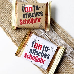 Könnte beinhalten: Zwei rechteckige Riegel in Goldfolie mit Papierbanderolen. Eine Banderole ist braun, die andere weiß. Beide tragen den Text "Ein fantastisches Schuljahr" in Schwarz und Rot sowie kleine Illustrationen von Bleistiften und einem lachenden Gesicht.