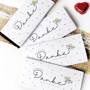 Può includere: Confezioni bianche di barrette di cioccolato con la parola "Danke" in corsivo, una grafica a margherita e piccoli accenti di stelle. Un involucro è parzialmente aperto, rivelando il cioccolato. Sono presenti anche una caramella rossa a forma di cuore e dei fiori.