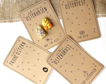 Puede incluir: Cuatro etiquetas de regalo de cartulina marrón con saludos de Pascua en alemán. Las etiquetas presentan pequeñas decoraciones con temática de Pascua, incluyendo un pollito, un conejito y una cesta de huevos. El texto en las etiquetas dice: "Einfach is FROHE OSTERN", "Liebe Gruße von OSTERHASEN", "Ein wundervolles OSTERFEST" y "Ein kleiner OSTERGRUSS".