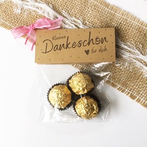 May include: A small cellophane bag containing three gold-wrapped chocolate candies. The bag is attached to a small brown tag with the words "Kleines Dankeschön für dich" printed in black script, tied with a pink ribbon.