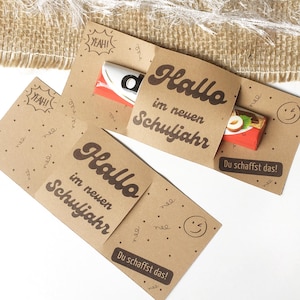 Könnte beinhalten: Geschenkkarten aus Kraftpapier mit dem Text "Hallo im neuen Schuljahr" und "Du schaffst das!" in Braun gedruckt. Die Karten haben einen kleinen Schokoriegel im Inneren. Der Hintergrund ist eine Jutestruktur.