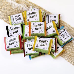 Könnte beinhalten: Eine Ansammlung kleiner rechteckiger Schokoriegel, eingewickelt in Papier mit Fußballmotiven wie Fußbällen, Gras und Tornetzen. Die Verpackungen zeigen deutschen Text wie Gelbe Karte, Halbzeit, Rote Karte und 2026 Zeit für einen Snack.