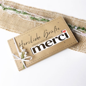 Peut inclure: Une boîte cadeau rectangulaire en papier brun portant le texte allemand Herzliche Grüße und ein riesengroßes merci avec une petite icône de cœur rouge. La boîte est ornée d'une ficelle blanche nouée en nœud et de petites fleurs blanches.