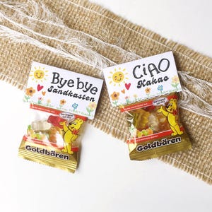 Könnte beinhalten: Zwei kleine durchsichtige Plastiktüten gefüllt mit Gummibärchen, jeweils mit einem weißen Papieraufhänger versehen. Ein Aufhänger zeigt Bye bye Sandkasten mit Sonne und Blumen, der andere CiAO Kakao. Beide Tüten tragen das Goldbären-Logo.