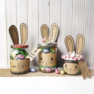 Peut inclure: Trois bocaux en verre décorés en lapins de Pâques. Chaque bocal a un couvercle différent et est rempli d'œufs de Pâques colorés et d'herbe décorative. Les bocaux ont des oreilles de lapin en papier et des étiquettes avec "Frohe Ostern".