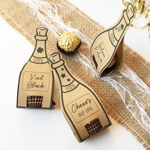 May include: Kraft paper champagne bottle-shaped gift boxes with the year 2026 printed on them. The boxes have different phrases in German, and are filled with chocolate. A gold-wrapped chocolate sits nearby.