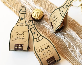 Può includere: Scatole regalo a forma di bottiglia di champagne in carta kraft con l'anno 2026 stampato sopra. Le scatole hanno frasi diverse in tedesco e sono riempite di cioccolato. Un cioccolatino dorato è vicino.