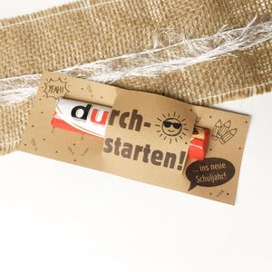 Op de afbeelding: Een rechthoekige bruine papieren wikkel om een chocoladereep. De wikkel bevat de tekst "durch-starten!" met een zonne-icoon met een zonnebril, het woord "YEAH!", potloodillustraties en een tekstballon met "... ins neue Schuljahr!".