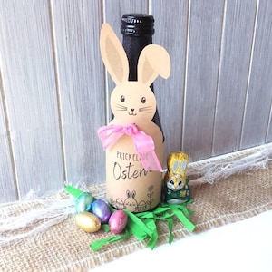 Puede incluir: Una botella de vino con temática de Pascua con un recorte en forma de conejito y una cinta rosa. La botella está rodeada de coloridos huevos de Pascua, hierba de papel verde y un conejito de chocolate. El texto "PRICKELNDE Ostern" es visible.