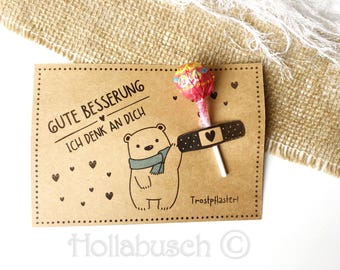 May include: A brown card with the text "Gute Besserung Ich denk an dich" and a small illustration of a bear holding a lollipop with a bandage on it. The text "Trostpflaster!" is at the bottom of the card.
