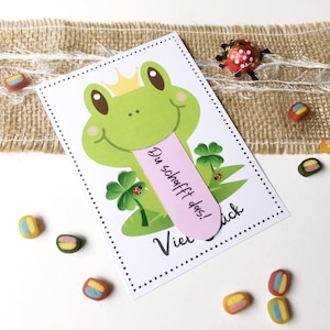 Könnte beinhalten: Eine Grußkarte mit einem Cartoon-Frosch mit Krone und rosa Zunge mit dem Text "Du schaffst das!". Die Karte enthält auch vierblättrige Kleeblätter und Marienkäfer. Bunte Süßigkeiten sind um die Karte herum verstreut.