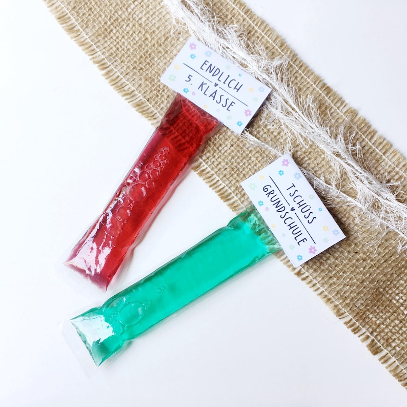 May include: Two frozen ice pops, one red and one green, in clear plastic packaging. Each pop has a small white tag with German text and floral accents. The pops are on a burlap and white surface.