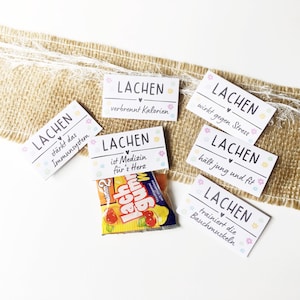 Könnte beinhalten: Eine Ansammlung weißer rechteckiger Karten mit dem Wort "LACHEN" und deutschen Sätzen, verziert mit Blumen- und Herzmotiven. Ein kleiner Beutel Gummibärchen ist ebenfalls zu sehen.