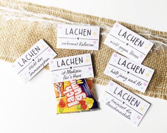May include: A collection of white rectangular cards with the word "LACHEN" printed on them, along with various German phrases. Some cards have floral and heart designs. A small bag of gummy candies is also visible.