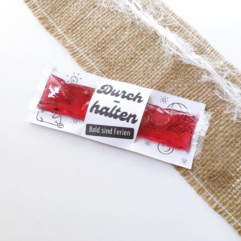 May include: A clear plastic package containing two red, rectangular, gel-like treats. The package is wrapped with a white paper band that has the words "Durchhalten Bald sind Ferien" printed on it in black. The background is a burlap material.