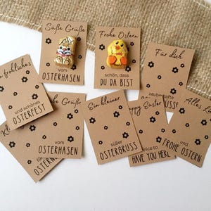 May include: A set of eight brown paper gift tags with black text and small black flower designs. The tags have various Easter greetings in German and English, including "Frohe Ostern", "Happy Easter", "Ein kleiner süßer Ostergruss", and "Alles Liebe". Two of the tags have small chocolate Easter figures attached.
