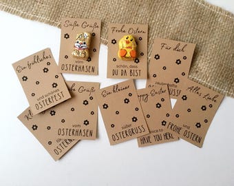 Puede incluir: Un conjunto de ocho etiquetas de regalo de papel marrón con texto negro y pequeños diseños de flores negras. Las etiquetas tienen varios saludos de Pascua en alemán e inglés, incluyendo "Frohe Ostern", "Happy Easter", "Ein kleiner süßer Ostergruss" y "Alles Liebe". Dos de las etiquetas tienen pequeñas figuras de Pascua de chocolate adjuntas.