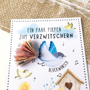 May include: A white card with a blue bird and a fan of money folded into the shape of a bird's tail. The card has the text "Ein paar piepen zum verzwitchern" and "Gl&uuml;ckwunsch" on it. There are also musical notes and small birds on the card.