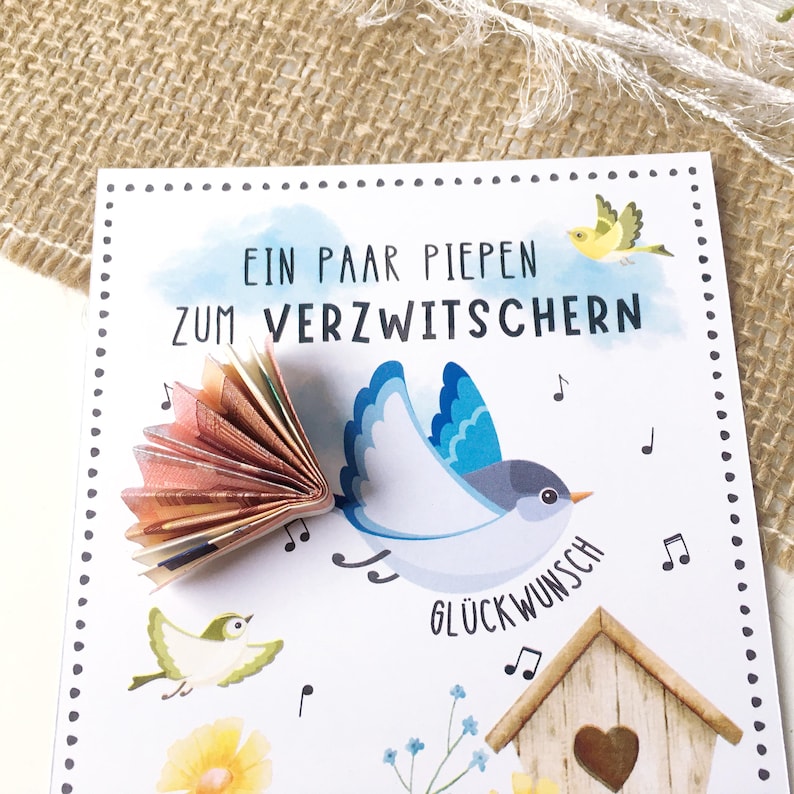 May include: A white card with a blue bird and a fan of money folded into the shape of a bird's tail. The card has the text "Ein paar piepen zum verzwitchern" and "Gl&uuml;ckwunsch" on it. There are also musical notes and small birds on the card.